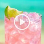 Closeup view of a pink drink in a glass of ice with a lime wedge garnish.