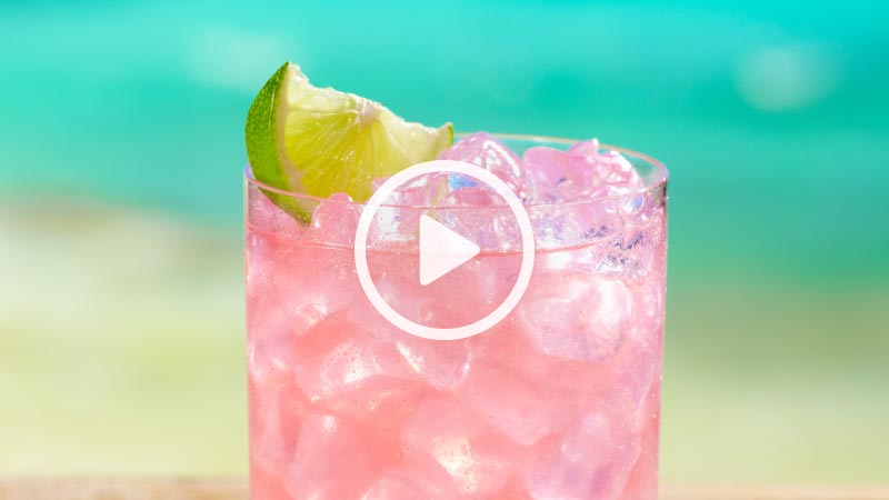 Closeup view of a pink drink in a glass of ice with a lime wedge garnish.